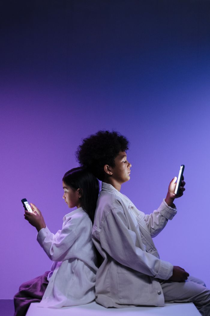 Two children using smartphones while sitting back to back in colorful lighting, highlighting digital engagement.